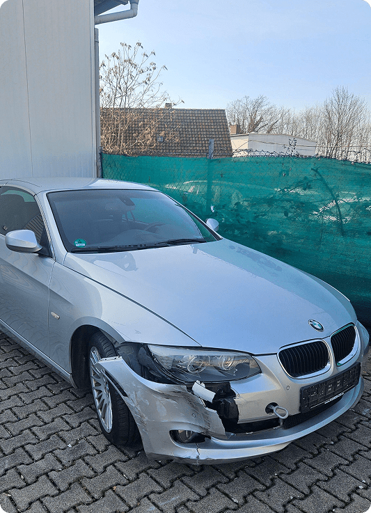 Silberner BMW mit beschädigter Frontstoßstange auf gepflastertem Parkplatz vor einem Gebäude.
